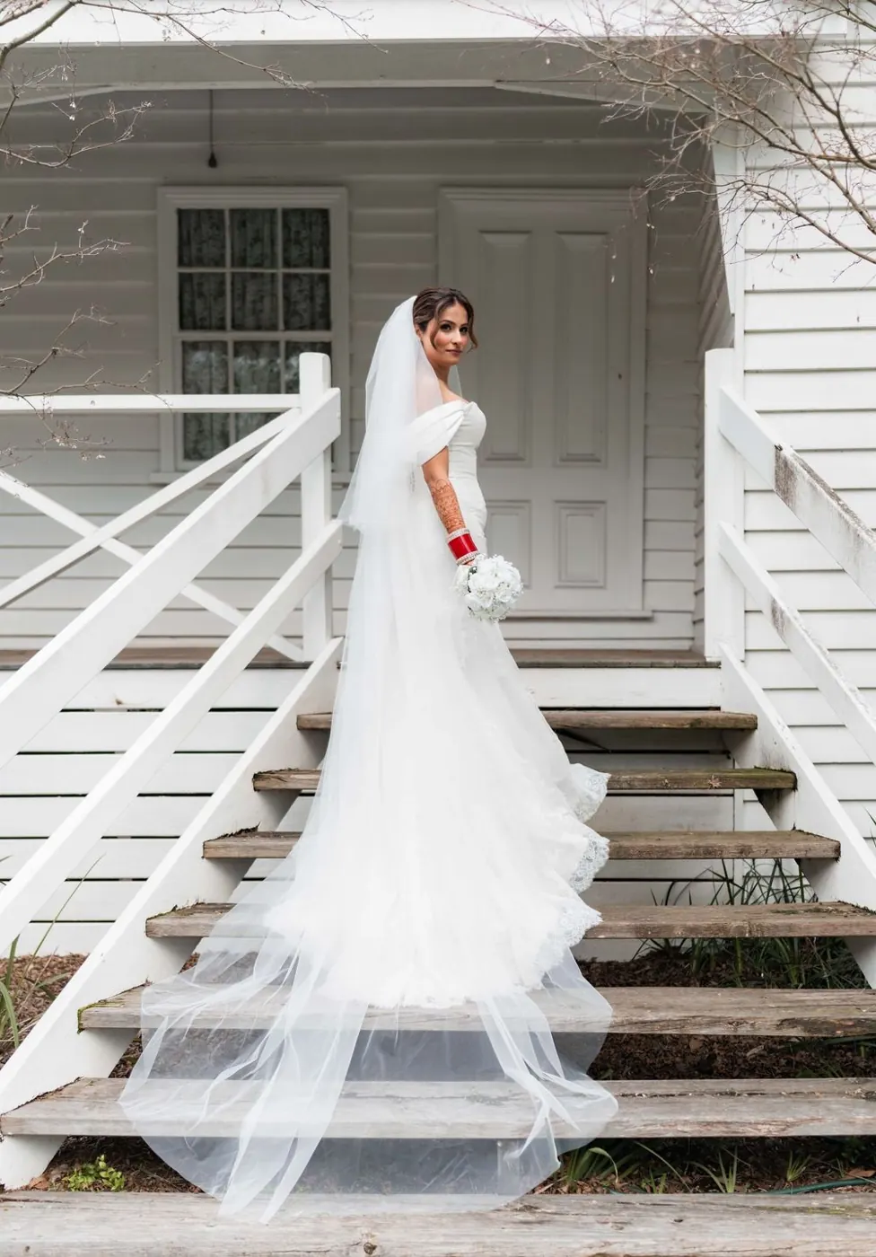 A bride in her wedding gown standing on the steps of a white country house.