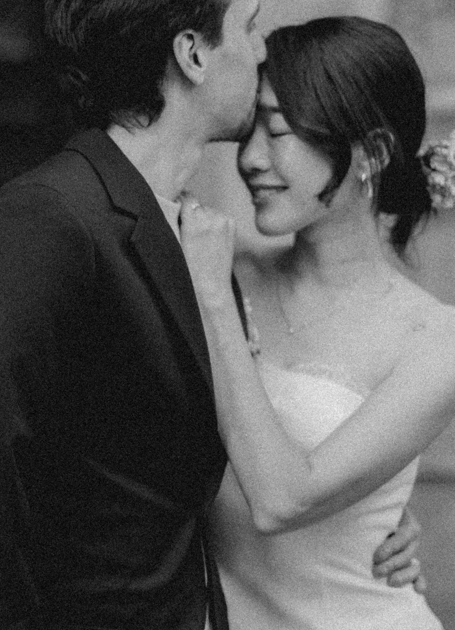 A groom kissing his smiling bride on the forehead, in black and white.