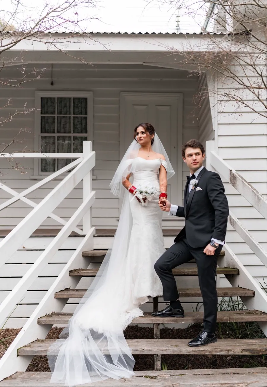 A bride and groom holding hands on the wooden steps of a white weatherboard cottage.