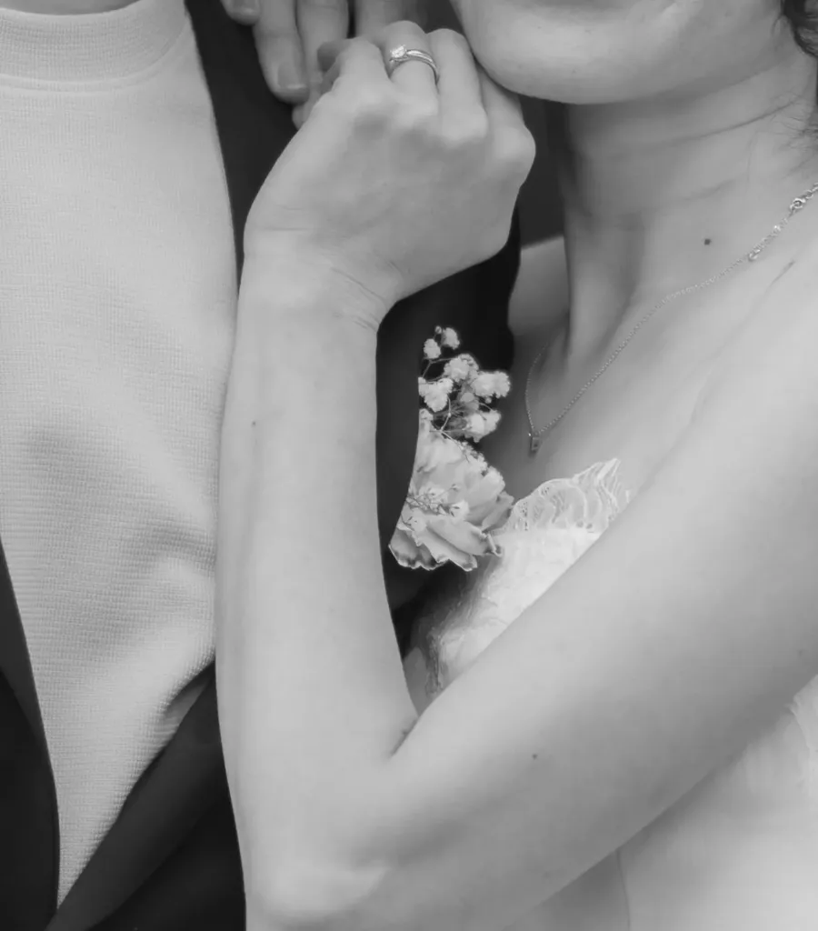 A close-up of a bride's hand and engagement ring resting on her groom's shoulder, in black and white.