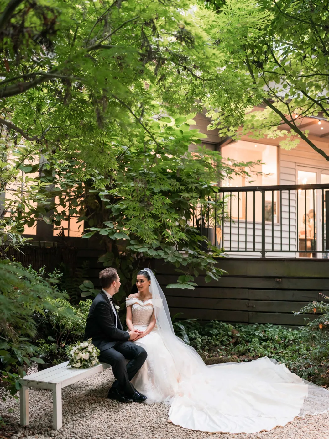 A bride and groom holding hands on a white bench in a leafy garden courtyard.